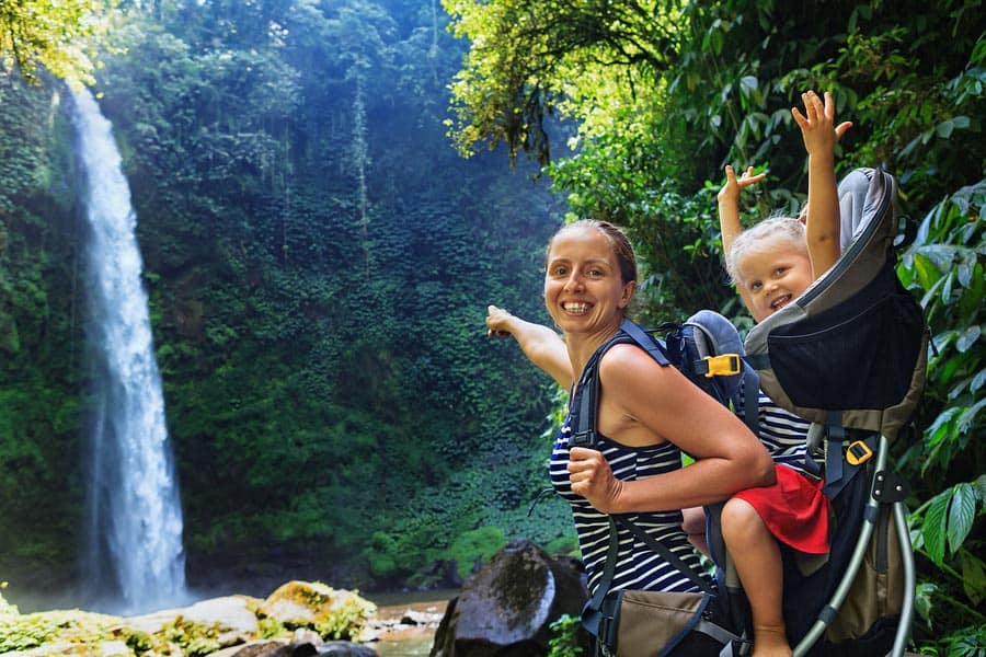 Hawaii vacations with kids. Young mother pointing to a waterfall with a baby in the backpack.